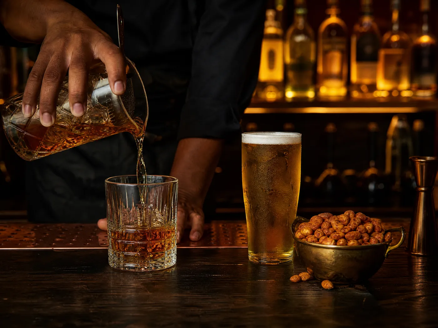 Whisky being poured beside a cold lager and masala peanuts at the bar.