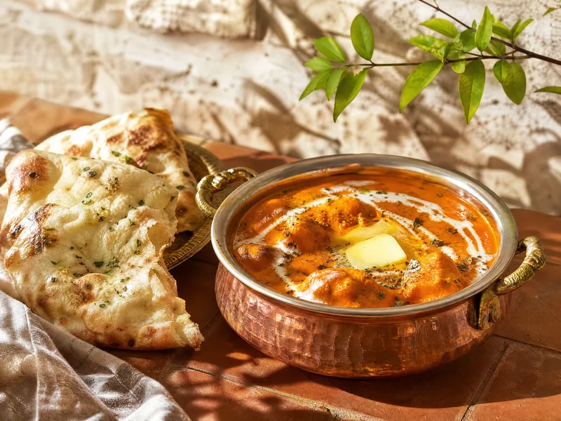 A copper pot of glossy orange butter chicken with a swirl of cream and a pat of butter, with naan.